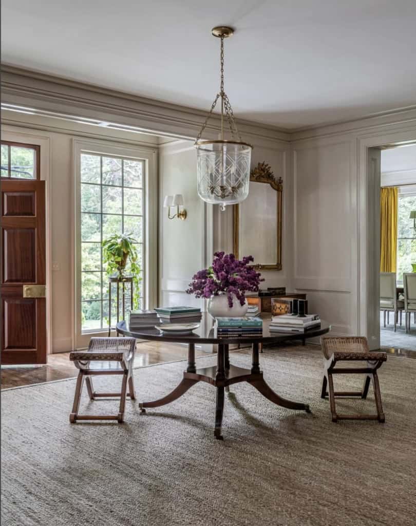 Elegant living room with a round wooden table, purple floral centerpiece, and large windows letting.